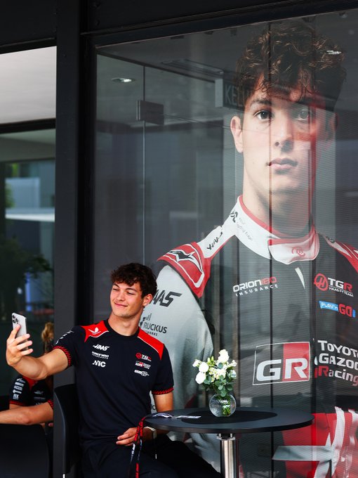 A young man in a black and red racing shirt taking a selfie, sitting in front of a large glass panel featuring a promotional image of himself in a Toyota Gazoo Racing suit.