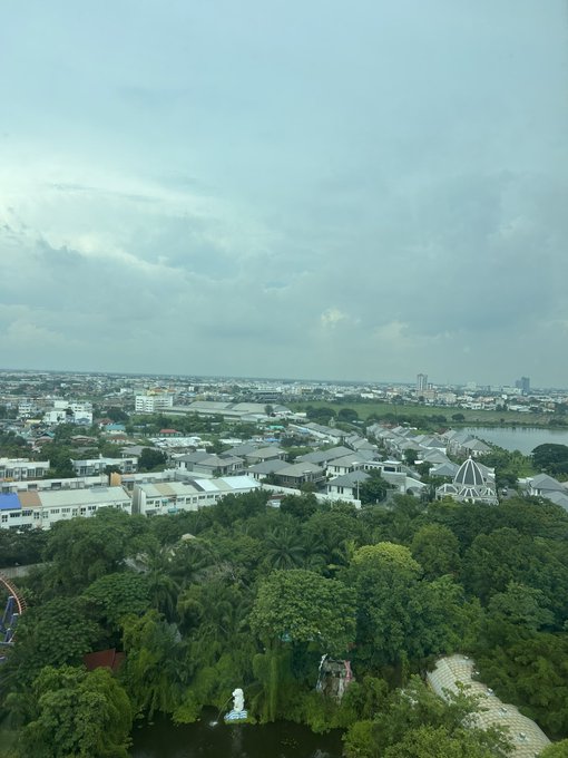 An elevated aerial view of a suburban cityscape with lush green trees and gardens in the foreground, residential buildings in the midground, and a cloudy sky above.