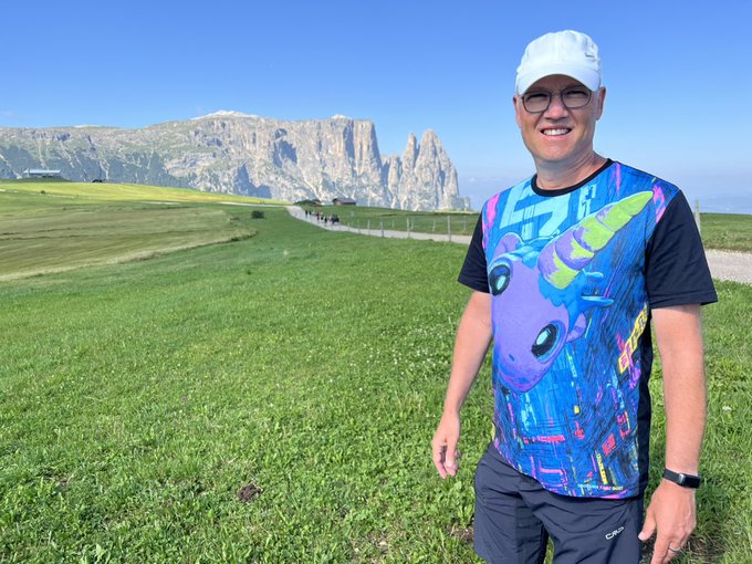 A smiling man in a white cap and colorful graphic t-shirt stands on a lush green alpine meadow with dramatic mountain peaks in the background. The scenic landscape symbolizes the post's message of "Proof of COMMUNITY $UFD," reflecting global unity and the beauty of diverse terrain that connects people everywhere.