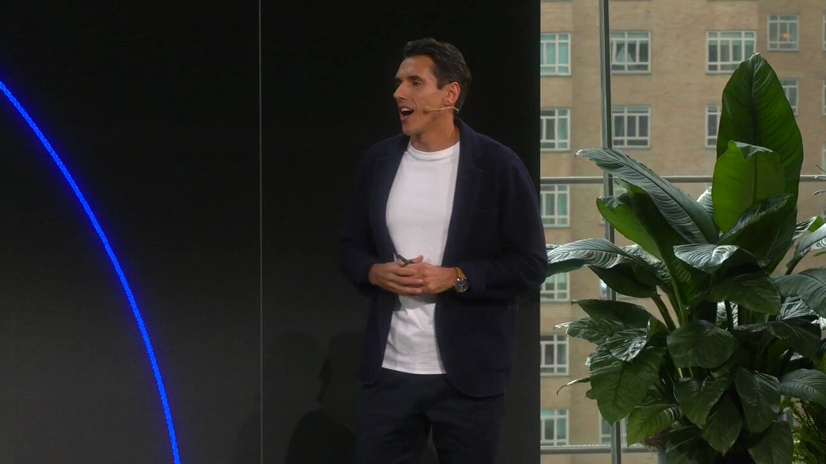 The image shows a man standing in front of a large window, giving a presentation or speech. The background features a green plant placed near the window, adding a touch of nature to the scene. There are also two blue lines visible on the wall behind the man, which could be part of an art installation or decoration.