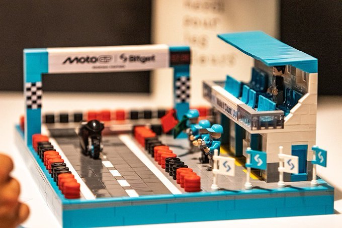 A Lego replica of a MotoGP pit lane with "MotoGP" and "Bitget" branding on the start banner, featuring a motorcycle, crew members, and numbered pit stalls. The miniature racing scene ties into the KCGI 2026 puzzle theme, where participants decode clues to identify Bitcoin, much like navigating the fast-paced, strategic world of crypto trading mirrored in motorsport.