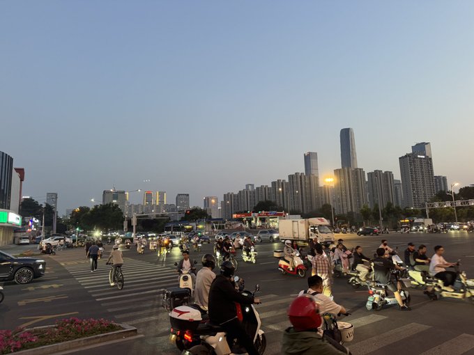The image shows a busy city street with many people riding motorcycles, mopeds, and bicycles. The scene takes place at dusk as the sun sets behind the buildings, casting long shadows across the street. There are numerous vehicles, including cars and trucks, parked or driving along the road. A few pedestrians can be seen walking on the sidewalks near the busy street.

The image also contains a post that reads "夜ラン かなり日が沈むのが遅くなってきました 仕事が終わっても明るいのは嬉しいですね #STEPN". This post seems to be related to the busy street scene, possibly discussing a specific event or location within the image.