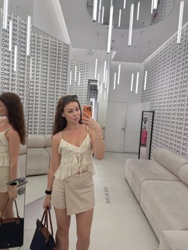 A woman wearing a cream-colored camisole top and beige shorts takes a mirror selfie inside a brightly lit store with patterned walls and modern lighting fixtures.