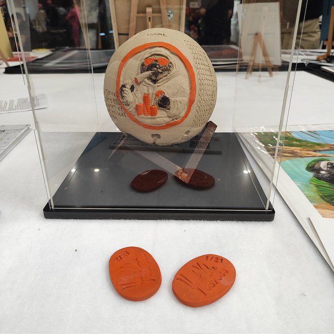 The image features a display case with two clay sculptures, including a baseball sculpture that has an orange face design. The sculptures are placed on a black base, which contrasts with their white color. In front of the display case, there are two small orange clay tokens, possibly representing the top five bidders in a competition or auction. The image also includes text that reads "Top 5 bidders each get one of these limited edition of 21 tokens. Link in first comment".