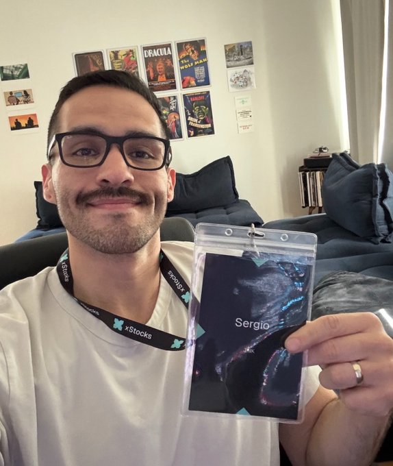 A smiling man with glasses wearing an xStocks lanyard, holding up a badge labeled 'Sergio' featuring an artistic portrait, with a wall of posters behind him including 'Dracula' and 'The Wild Robot'.