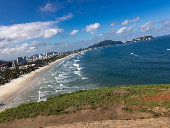 A scenic aerial view of a long sandy beach with waves, a coastal city skyline, and mountains in the distance under a partly cloudy blue sky.