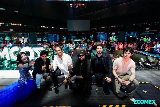 Group photo of six people posing together at what appears to be a Zoomex crypto event, with a woman in a blue dress on the left and men in various casual outfits, with the ZOOMEX venue and branding visible in the background.