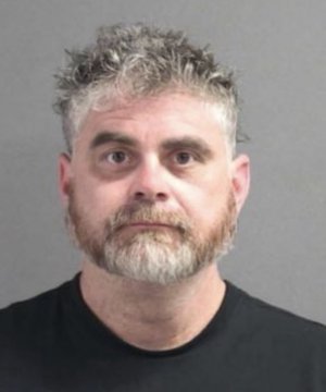 A man with graying curly hair and a beard appears in what looks like a mugshot photo, wearing a black shirt against a plain gray background. The image doesn't appear to have any visible text, numbers, or names, and there's no clear connection to the "$70K January / $70K August" reference in the accompanying post.