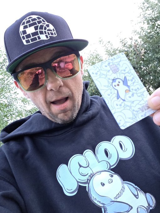 A man wearing an igloo-logo cap, reflective sunglasses, and a black hoodie featuring a v-tuber penguin character holds up a credit card decorated with a similar cute penguin design.