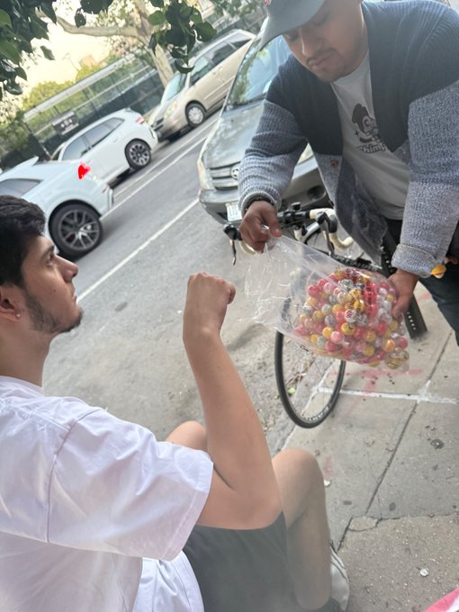 A young man in a white shirt sits on the street as a vendor in a gray cardigan offers him a clear plastic bag filled with colorful round candies.
