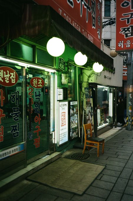 A nighttime street scene in Korea featuring traditional Korean restaurant storefronts with red signage displaying Korean characters, glowing round lanterns, menu boards, and a wooden chair placed outside on the sidewalk.