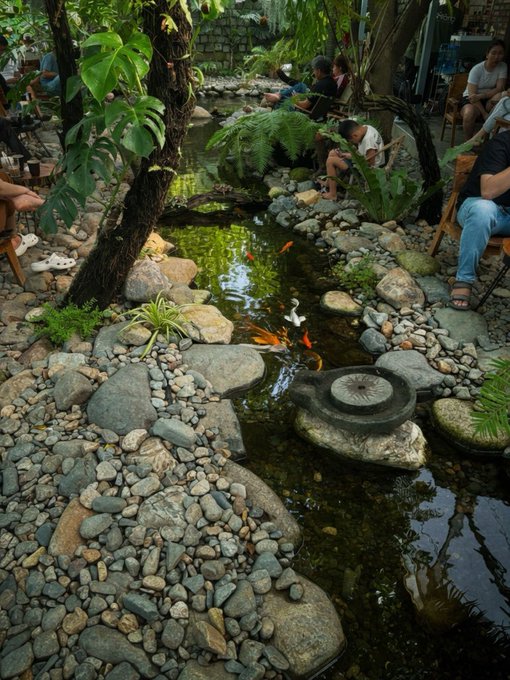 A tranquil outdoor café features a koi pond surrounded by smooth river stones, lush greenery, and patrons relaxing on wooden chairs, with the water providing a calming focus away from the workday grind.

The image's peaceful pond scene pairs nicely with the post's encouraging message to coworkers about receiving their pay—offering a visual moment of zen to counter the anticipation and hope for financial relief.