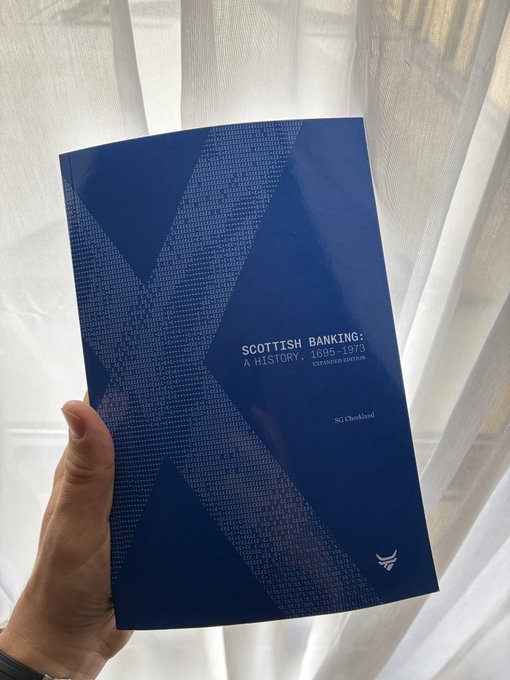 A hand holding a blue book titled 'SCOTTISH BANKING: A HISTORY, 1695-1973, REVISED EDITION' by S.G. Checkland, photographed against white curtains.