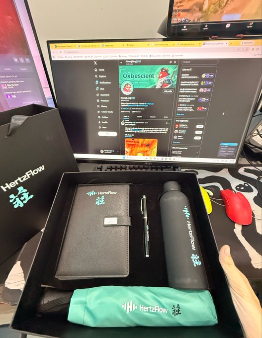 An unboxing of a HertzFlow branded gift set containing a notebook, pen, water bottle, and drawstring pouch, placed on a desk with a monitor displaying Twitter/X in the background.