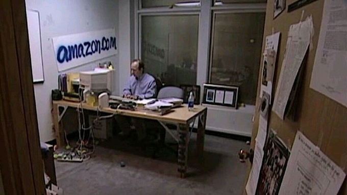 A vintage photograph shows a man working at a desk in a small, cluttered office with a whiteboard reading 'amazon.com' behind him, recognizable as Jeff Bezos in Amazon's early days.