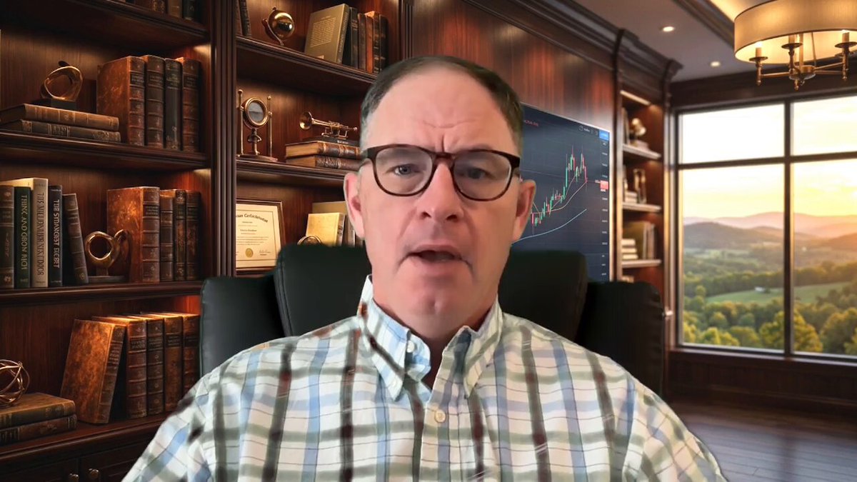 The image shows a man sitting down with a serious expression, possibly presenting or discussing something at a conference. He appears to be wearing glasses and talking into a microphone, indicating that he could be giving a presentation or speech. The room features a large window in the background, which provides an outdoor view of green trees outside. There is also a bookcase filled with numerous books visible in the image, adding an element of professionalism to the setting.