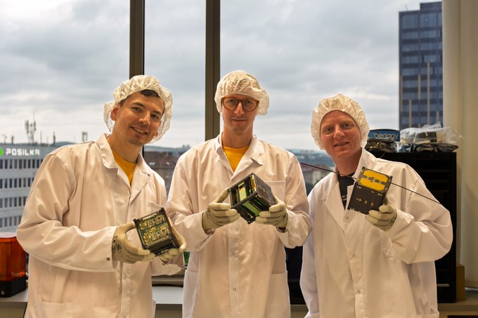 Three men in white lab coats and hairnets posing with electronic hardware/components, likely a satellite or aerospace project, with a 'PostNord' building visible through the window behind them.