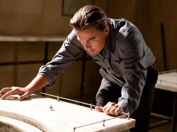 A man in a gray shirt leans over a table examining what appears to be a model or architectural layout. The image relates to the post by capturing a moment of intense scrutiny and analysis, mirroring how investors carefully study stocks and memes to find patterns or connections—suggesting both require a keen eye for detail and a willingness to lean in closer for a better look.