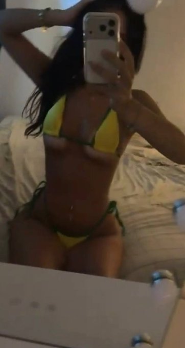 A mirror selfie of a woman in a yellow and green bikini sitting on a bed, covering her face with her phone.