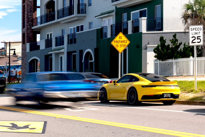 A yellow Porsche and a blurred blue vehicle on a street with a 'BE PREPARED TO STOP' sign and a 25 MPH speed limit sign.