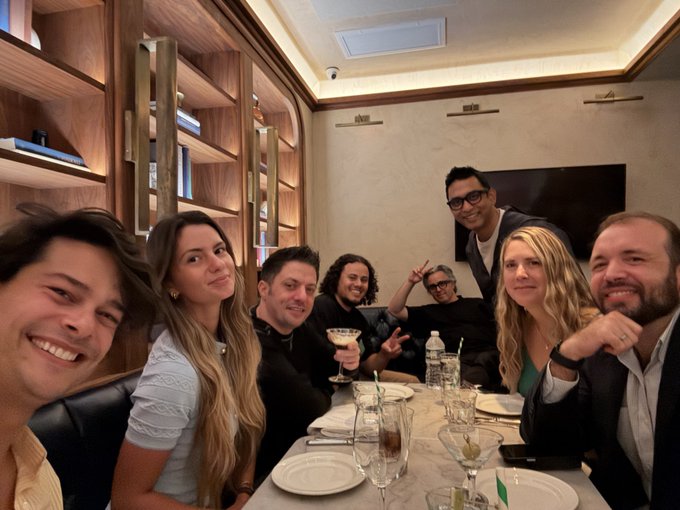 A group selfie of seven people smiling at a restaurant table, with elegant wooden shelving and ambient lighting visible in the background.
