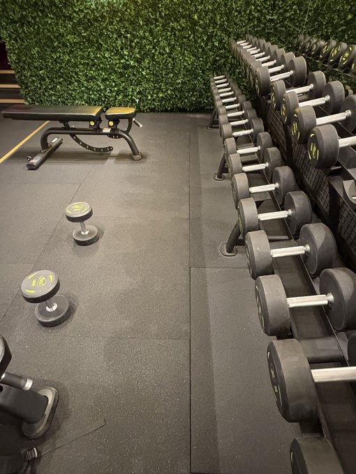 A gym workout area featuring a rack of black dumbbells lined up on the right, a flat weight bench in the background, and two smaller dumbbells resting on the rubber floor — a fitting scene for the post's message about staying consistent and grinding through training as the date changes.