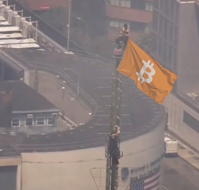 A person climbs a tall flagpole to raise an orange Bitcoin (₿) flag against an urban backdrop, symbolically marking the cryptocurrency's prominence as referenced by the #Bitcoin hashtag.
