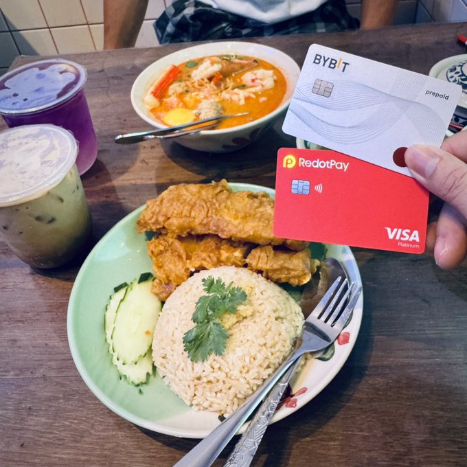A hand holds two prepaid cards—a white 'BYBIT prepaid' card and a red 'RedotPay VISA Platinum' card—above a wooden table set with a plate of chicken and rice, a bowl of noodle soup, and two iced drinks.
