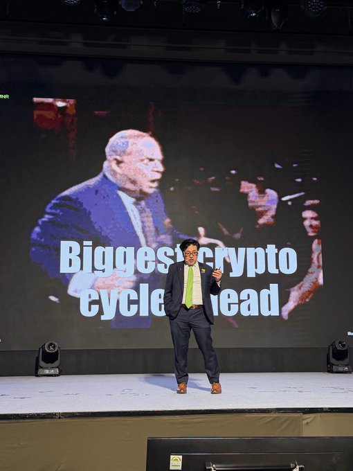 A man in a suit and tie stands on stage in front of a large screen displaying an older man and the text "Biggest crypto cycle ahead," suggesting the speaker is delivering a presentation about an upcoming cryptocurrency bull market, aligning with the enthusiastic tone of "Yas."