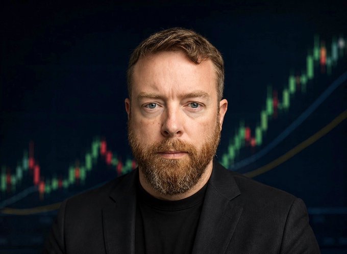 A portrait of a bearded man in a dark blazer looking at the camera, with a green and red stock market candlestick chart in the background.