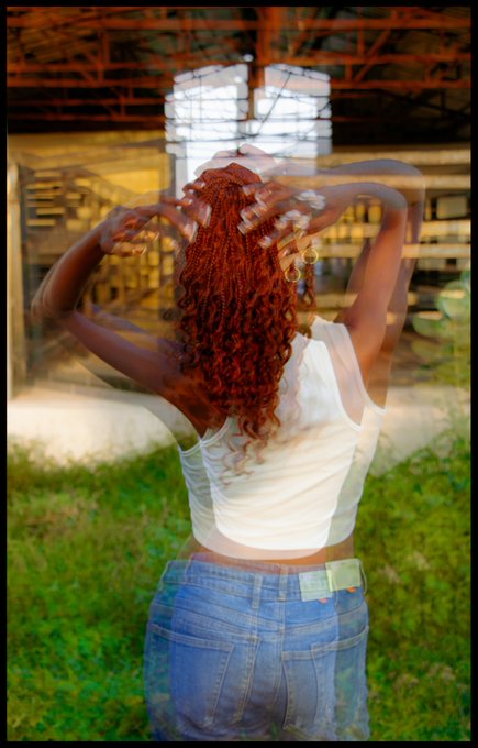 A double-exposed photo showing a woman with curly red hair in a white tank top and jeans, her hands raised, blended with an industrial structure and green grass.
