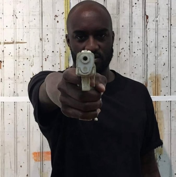 A Black man in a dark t-shirt points a small white handgun directly at the camera.