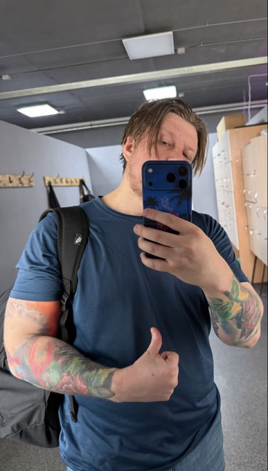 A mirror selfie of a young man in a blue t-shirt taking a photo with a dark smartphone, displaying colorful sleeve tattoos on his right arm and carrying a black backpack.