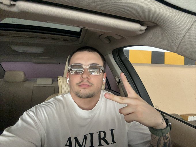 Selfie of a man inside a car wearing reflective sunglasses, a white "AMIRI" t-shirt, and AirPods, making a peace sign with a tattooed arm.