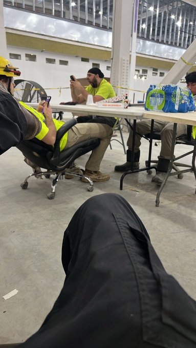 Construction workers in yellow safety vests sitting at a folding table, with someone in the foreground looking at their phone.