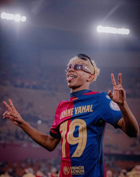 A soccer player wearing a Barcelona jersey with "RAHIM VAMAL" and number 19 celebrating on a stadium field, wearing sunglasses and making peace signs.