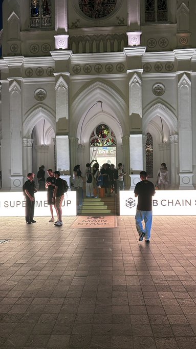 An interior view of a grand, cathedral-like building with illuminated signs for 'SUPERME UP' and 'BNB CHAIN' at an event.