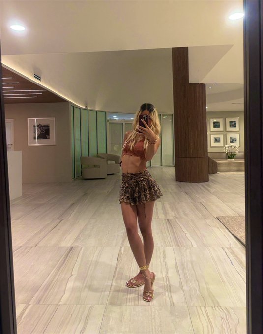 A mirror selfie of a woman in a reddish-brown top and animal-print mini skirt, taking a photo with her phone in a modern, spacious room with marble floors.
