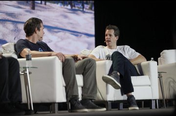 Two men seated in white chairs on a stage during what appears to be a panel discussion or interview.