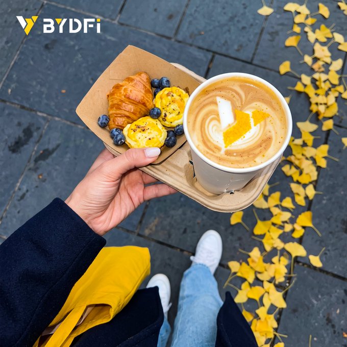 A first-person view of a hand holding a coffee cup and a paper plate with a croissant, blueberries, and pastries, with the BYDFi logo in the upper left corner and autumn leaves scattered on the ground.