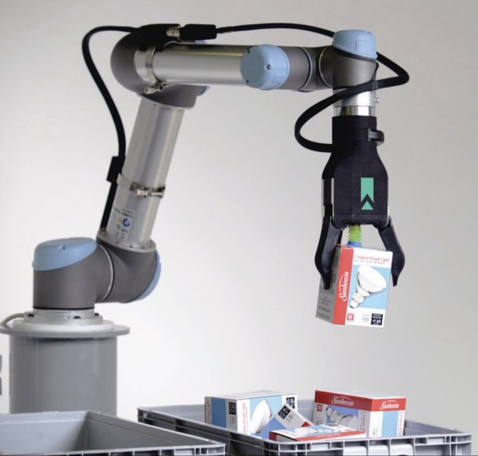A Universal Robots robotic arm with a gripper handling Sunbeam LED light bulb packages above a bin of similar products.