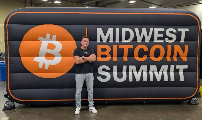 A man stands with crossed arms in front of a large banner for the 'MIDWEST BITCOIN SUMMIT' featuring a Bitcoin logo.