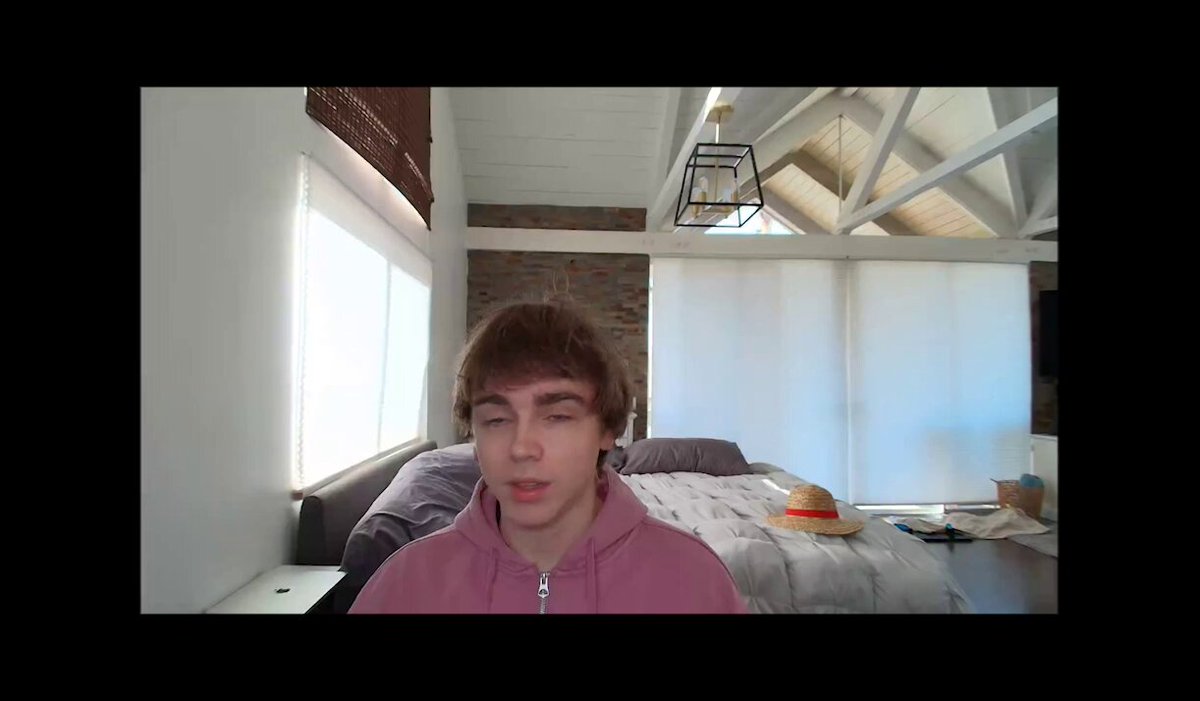 The image shows a young man wearing a pink hoodie sitting at a desk with his eyes closed while looking directly into the camera. The background of the image features a bedroom setting, which includes a bed and a chair.