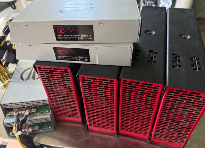 A collection of Obelisk cryptocurrency mining equipment is shown, featuring silver metal units on top and black/red units below with visible power cables.