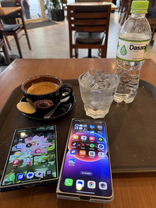A casual café scene showing a brown tray holding an espresso cup, a glass of ice water, and a bottle of Dasani water, alongside two smartphones (one displaying a floral wallpaper, the other showing various app icons).