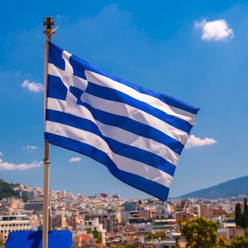 A large blue and white striped Greek flag waving in the wind against a blue sky, with a cityscape and hills visible in the background.