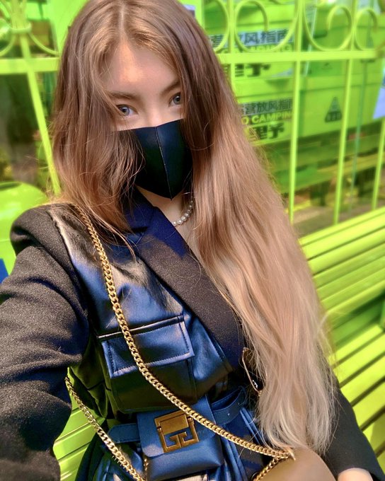 A young woman with long wavy blonde hair wearing a black face mask and a blue leather Fendi bag, standing in front of a bright green bench.