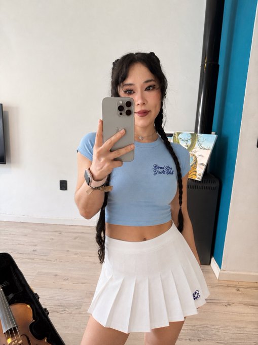 A mirror selfie of a young woman with long dark braided hair wearing a light blue crop top and white pleated tennis skirt, holding an iPhone in a room with a violin visible.