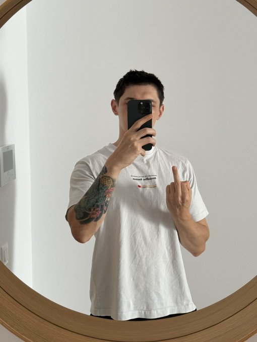 A person with a tattooed arm takes a mirror selfie while wearing a white shirt with small text, holding a phone in front of their face and making a hand gesture.