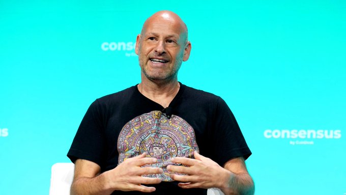 A bald man with a goatee, wearing a black t-shirt featuring a colorful Mayan-style calendar design, smiles while speaking on a stage at the "consensus" conference. The image relates to the post as it likely depicts Joseph Lubin, founder of ConsenSys, the company spinning off MetaMask into its own entity.